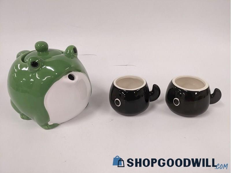 a product photo of a teapot shaped like a very round green frog. the spout is the frog's mouth, open in a small O. next to the teapot are two black tadpole mugs.