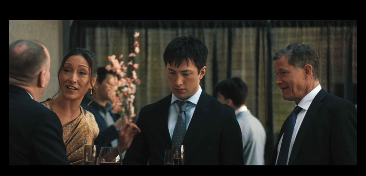 shane stands in a nicely dress room, flanked by his parents. his mom, on the left, is talking to the owner of the montreal metros, who has his back to the camera. yuna is wearing a shiny, gold dress. shane is wearing a black suit jacket, eggshell blue shirt, and blue-gray tie with a diagonal grid pattern. his dad is wearing a standard suit with a black suit jacket and tie with white shirt.