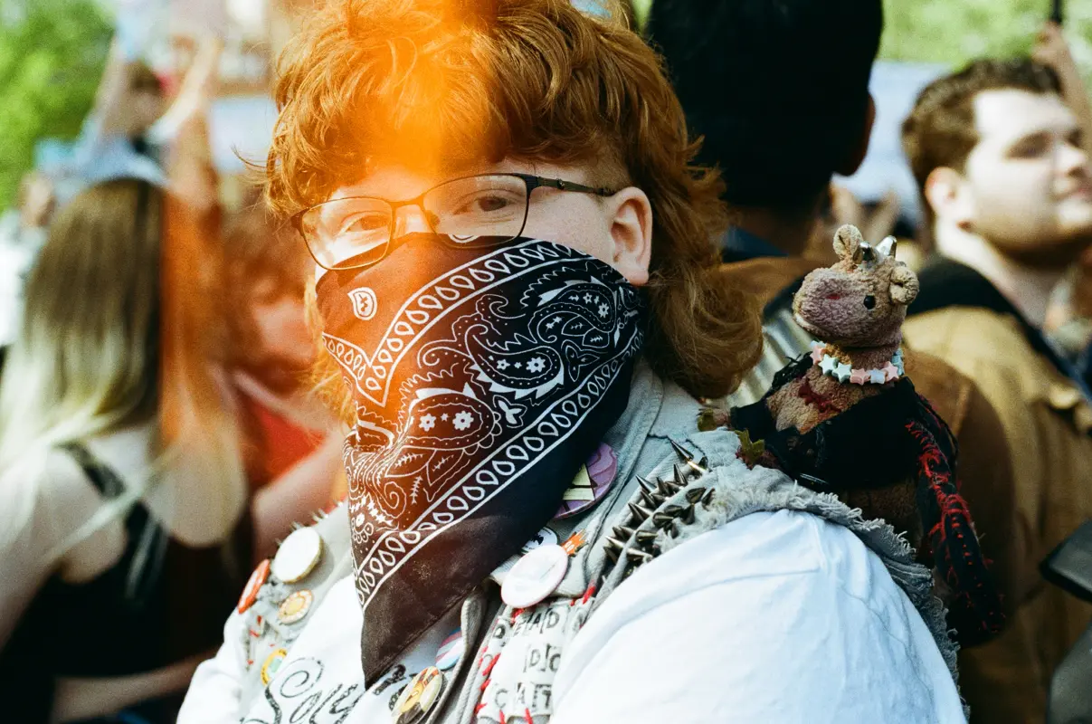 a person wearing a scarf over their nose and mouth is looking at the camera, on their shoulder a toy unicorn with a kandi necklace.