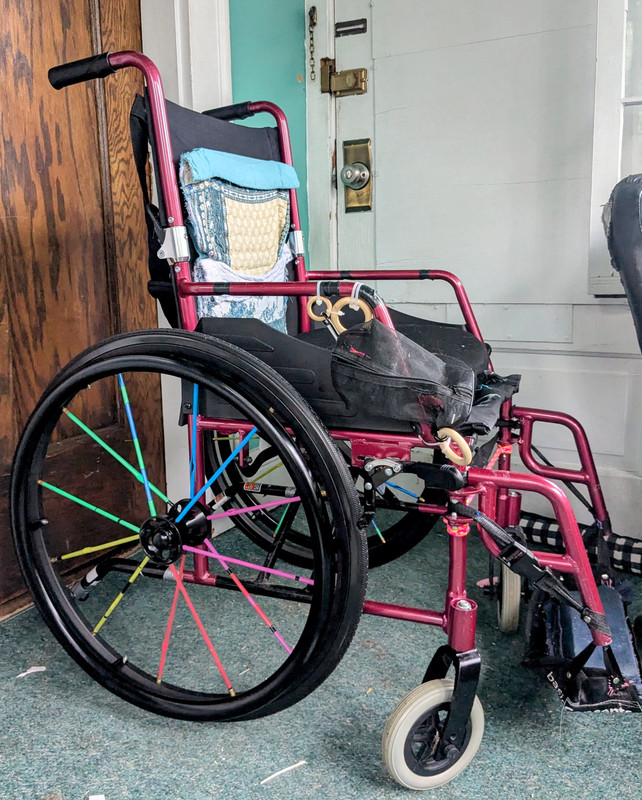 A black and burgundy wheelchair that has been modified. It has a teal paisley backrest; larger black wheels with rainbow spoke decorations; a home-made footplate supported by nylon straps; and some attachmrnts zip-tied to the arms.