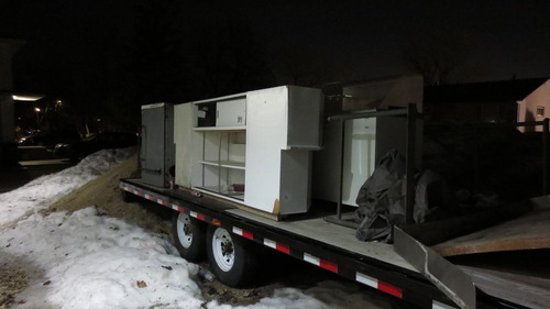 A flatbed trailer outside on a mix of gravel and snow, at night. On it sits a series of counters, stove tops and other kitchen equipment.