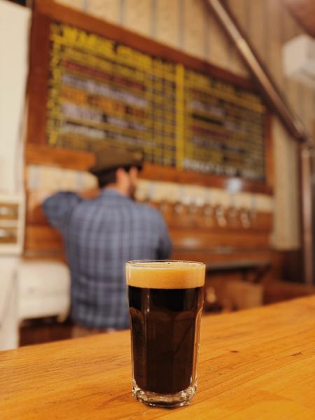Vaso de cerveza negra frente a la barra y la pizarra de cervezas de Cervecería Rural