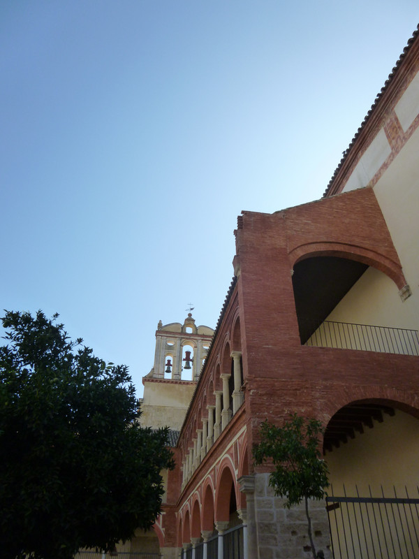 Red and white painted arches in a courtyard behind a church.  At the top of the picture, two out of three church bells can be seen.  They are also in arches.  In the foreground are trees.  One, on the right, is very small.