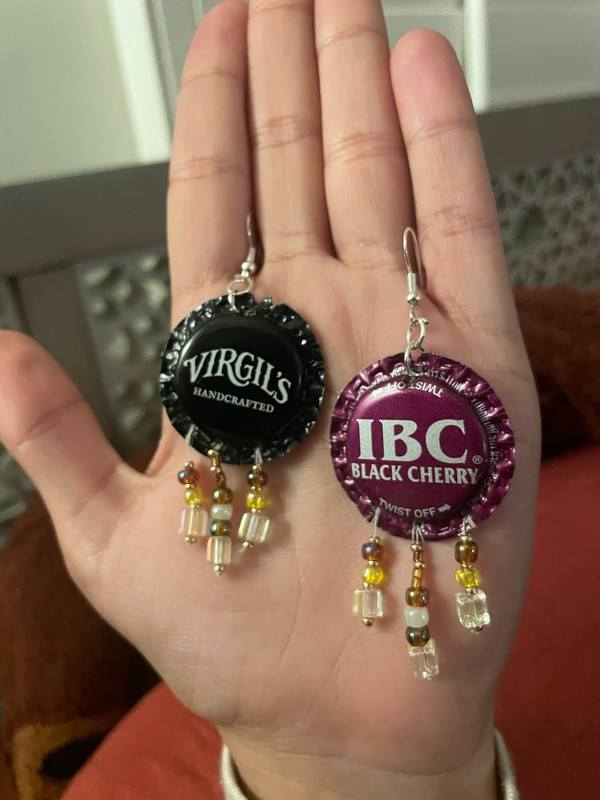 a photo of a hand holding a pair of bottlecap earrings. one is a black virgil's soda bottlecap and the other is a purple IBC black cherry soda bottlecap, both with three strings of light yellow/gold/white beads hanging off of them