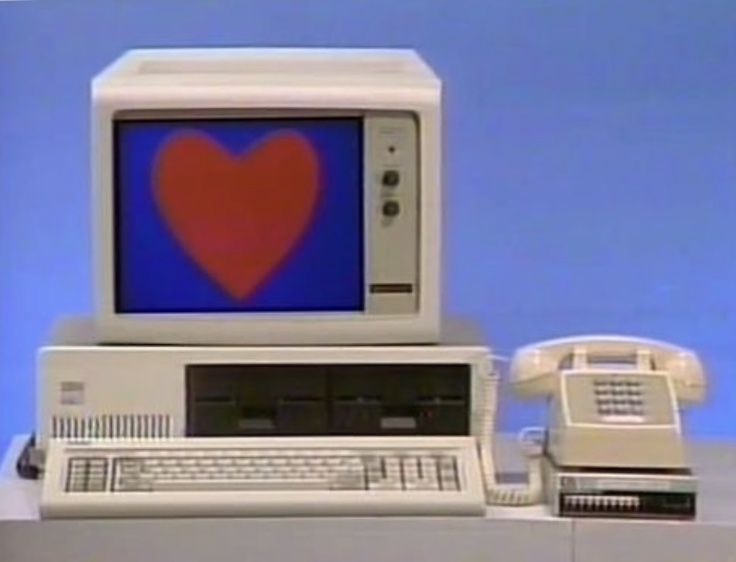 an old computer with a heart on the screen. the computer is next to a telephone