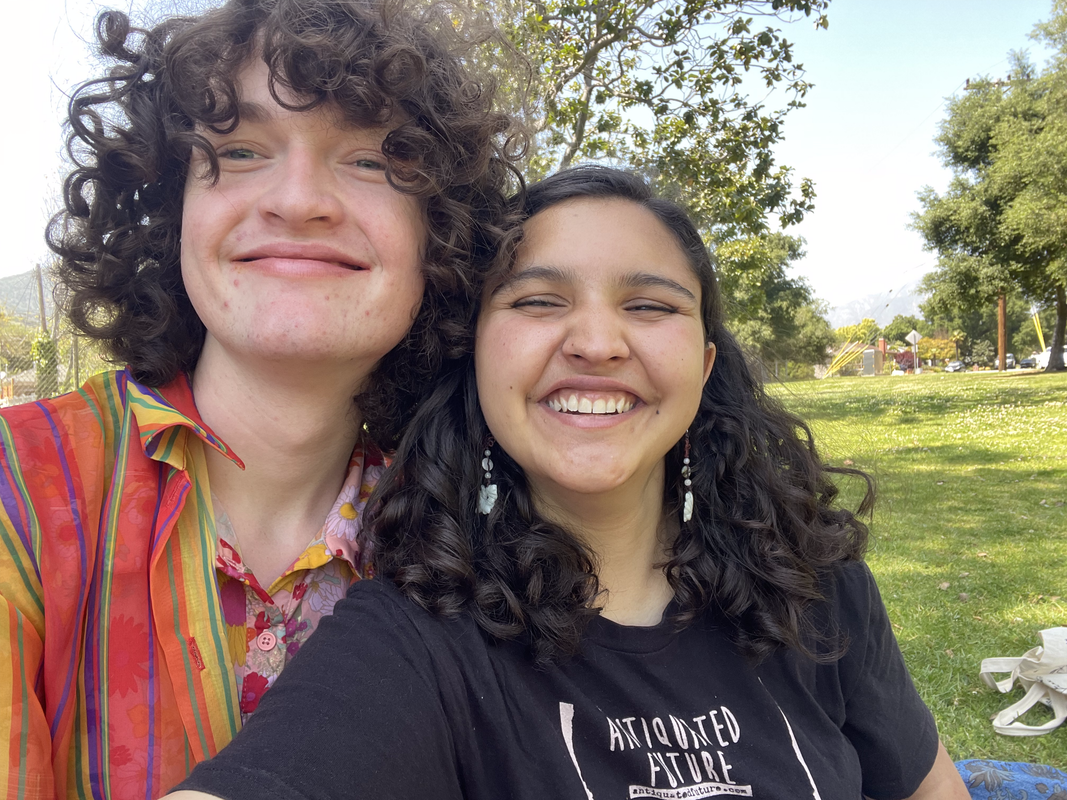 a photo of two people smiling-- one who is white with very curly light brown hair and bangs, wearing a colorful top and button up and another who is brown and has dark brown, long curly dark hair and is wearing black-- my girlfriend and i, respectively. you can see a park behind us.
