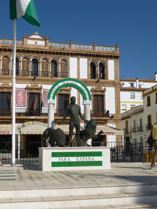 Statue of Hercules holding 2 lions.  This is a reference to the mythological tale of Hercules being the founder of Andalucia.  Behind him is a green-white-green arch held up by two pillars.  Across the arch it says "Dominator Hercules Fundator".  At the front of the statue it says "Andalucia Por Si Para Espana Y La Humanidad" (Andalucia for itself, for Spain and humanity, I think).

The statue is in front of an 1800 style building, white stone with brown edges.

In front of the statue, cut off by the top of the photo is a green-white-green flat of Andalucia on a flag pole.