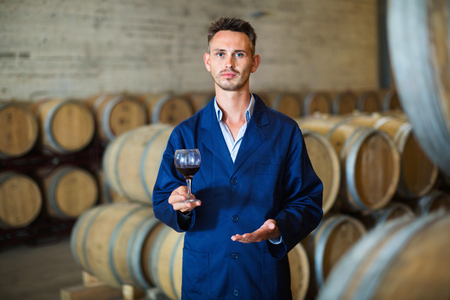 64918503-smiling-male-efficient-winemaker-in-uniform-having-glass-of-wine-in-hands-in-cellar