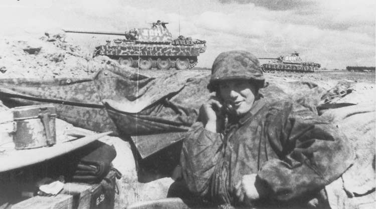 Un Panzergrenadier recibiendo instrucciones. Al fondo varios carros medios Pz.Kpfw.V Panther se dirigen al combate