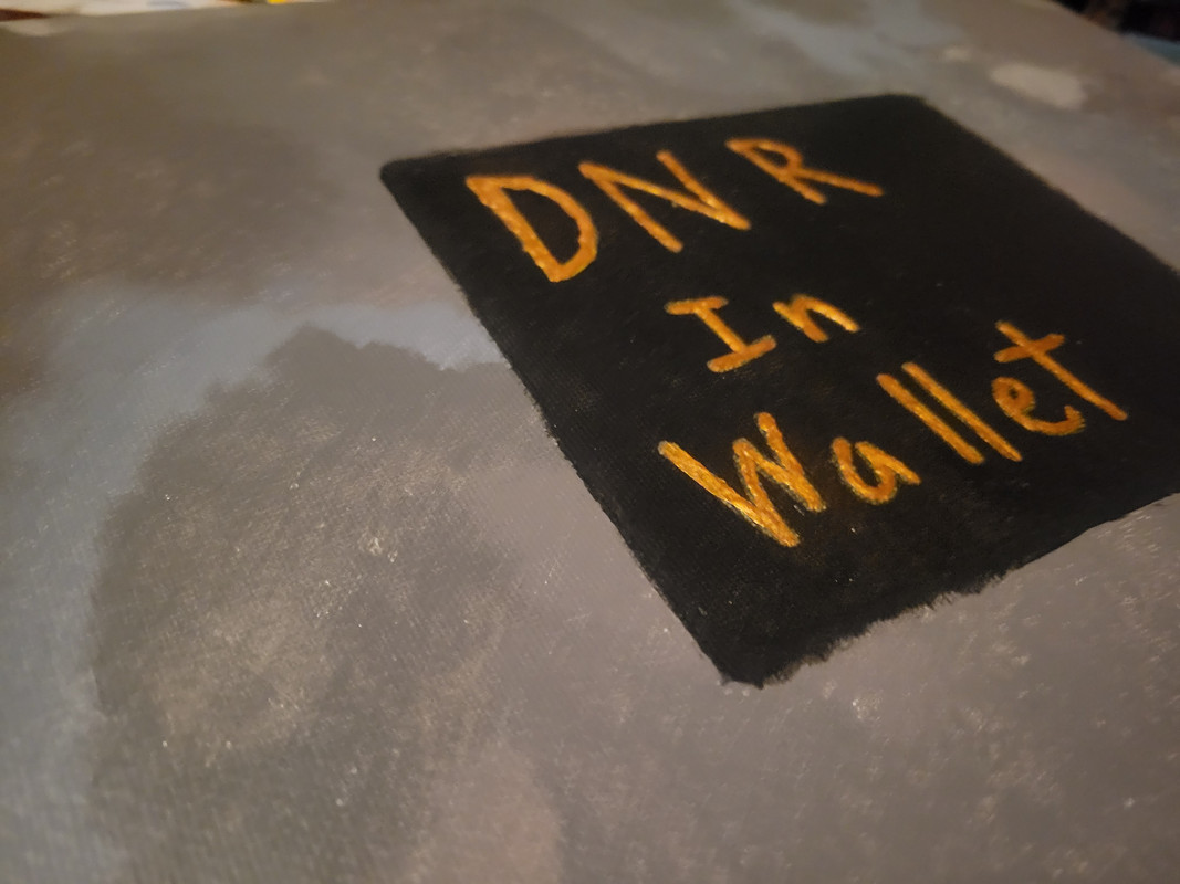 A close up picture of an acrylic painting of a black square on a gray background The black sqaure has gold text on it that reads DNR In Wallet yhe picture is taken from the lower left corner of the painging with the painting and the lower right corner of the black square is cut off because of this