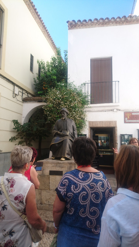 Metal statue of Maimonides.  He is wearing a cloth cap and a robe.  The metal is a dark brown, but there are golden glints where it is worn.  This is particularly strong on the feet of Maimonides, the bottom edge of his beard and robe, and around the book he is holding.