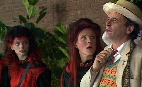 Screencap from Paradise Towers: the Doctor stares into the middle distance, while two Red Kangs (young women wearing red and with black- and red-dyed hair) look at him in bemusement.