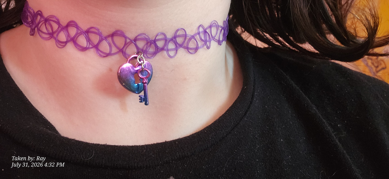 a close up of the choker I'm wearing with the Lisa frank shirt. The black shirt collar is visible. he choker is a purple tattoo choker with a dangly heart shaped lock with a dangly little key, both the charms are a pink purple blue gradient so it's accidental bi pride shit i love it