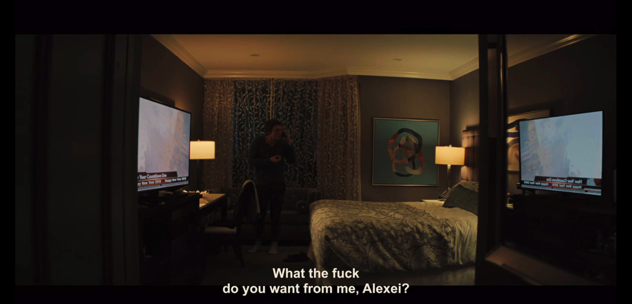 ilya in shadow standing in a hotel room lit only by the tv and lamp. he's wearing a long-sleeved gray shirt, black pants, and white socks.