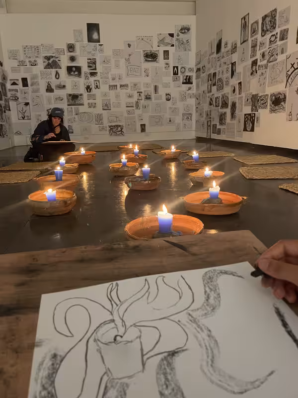 a photo of a piece of artwork of a burning candle on the lap of a person, with a room spanning out in front of them that has little mats with candles on the floor and lots of drawings posted on the walls. it's an interactive art gallery exhibit.
