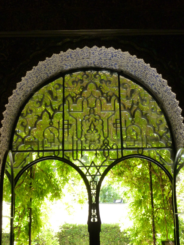Ornate wrought iron archway, looking almost caligraphic.  The foreground is shadowed, and you can see the carved stonework of the ceiling and wall that the arch fits into.  Outside are verdant green leaves on trees.