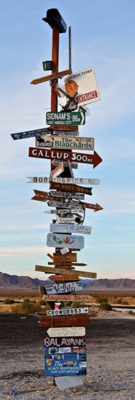 An insanely tall wooden poll with very, very many signs of various sizes, shapes, colours, and words. There are a few arrow signs pointing in various directions, and a mailbox on the very top. In the background is a light blue sky with faint clouds, and mountains to the bottom of the image.