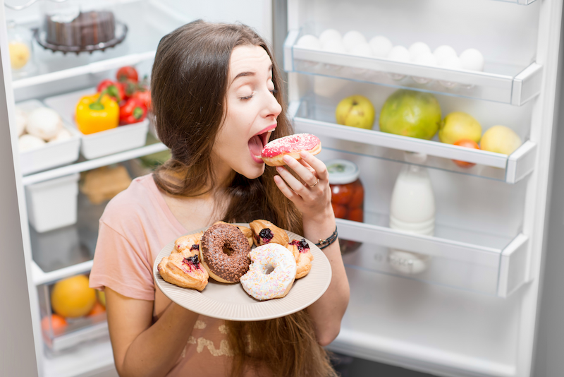 Woman taking a bite of a doughnut in front of a fridge 