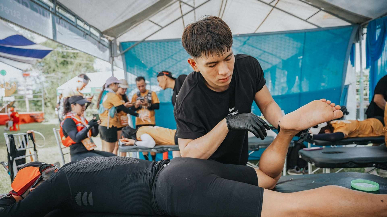 Một kỹ thuật viên đang Massage đùi cho một vận động viên tập HYROX tại khu vực phục hồi thể chất ở ngoài trời. Vận động viên đang nằm trên bàn massage. Ảnh minh họa cho kỹ thuật Sport Massage của Active Fox, phục hồi cơ bắp, trị liệu đau chân và tầm quan trọng của việc chăm sóc cơ thể sau khi tập luyện.
