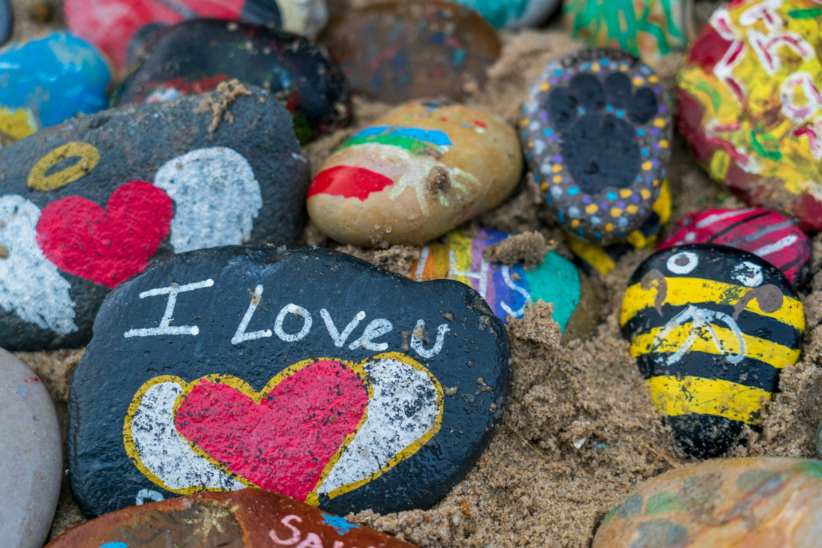 Painted rocks on sand, including designs like a bee, a heart with wings, a footprint, and the message “I love you”—part of a calming summer art activity for stress relief and creative expression..