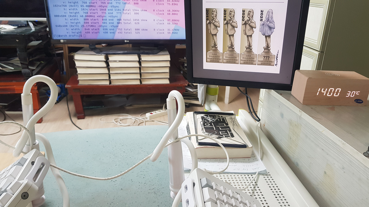 The left-hand table and footstool are unchanged, but the right-hand table has been replaced by a much smaller white steel desk extending from a vertical pole. The computer housing is now to the right of the steel desk, and the vertical monitor is suspended from the same pole as the new right-hand desk.