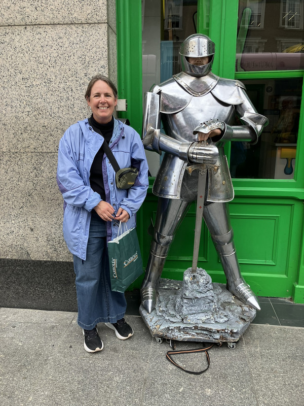 Dr. Valerie Couture, a smiling woman with pull-backed brown hair, standing outdoors next to a full suit of silver armor holding a sword. She is wearing a light blue jacket over a dark top, wide-leg blue jeans, black sneakers, and a small crossbody bag, while carrying a green shopping bag outside a shop with a bright green storefront.