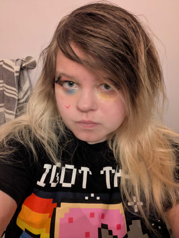 Selfie of the owner of the website They are white, wearing big eyeliner. blue, yellow, white and red eyeshadow with pink hues and glitter. They have Simple Scene hair that is brown at the roots and becomes a barbie blonde further down. They are wearing a nyan cat shirt.