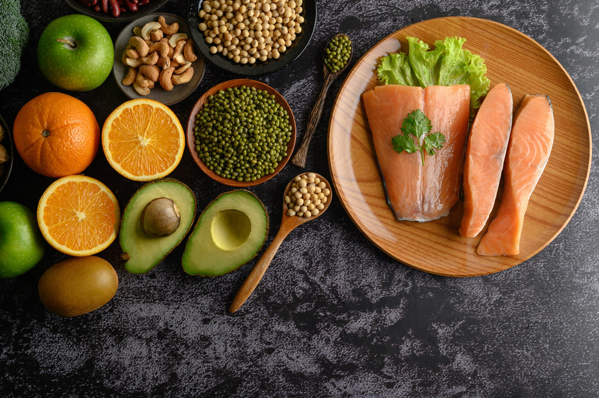 Un assortimento di cibi freschi e sani disposti su un piano scuro, tra cui salmone, avocado, frutta e verdura.