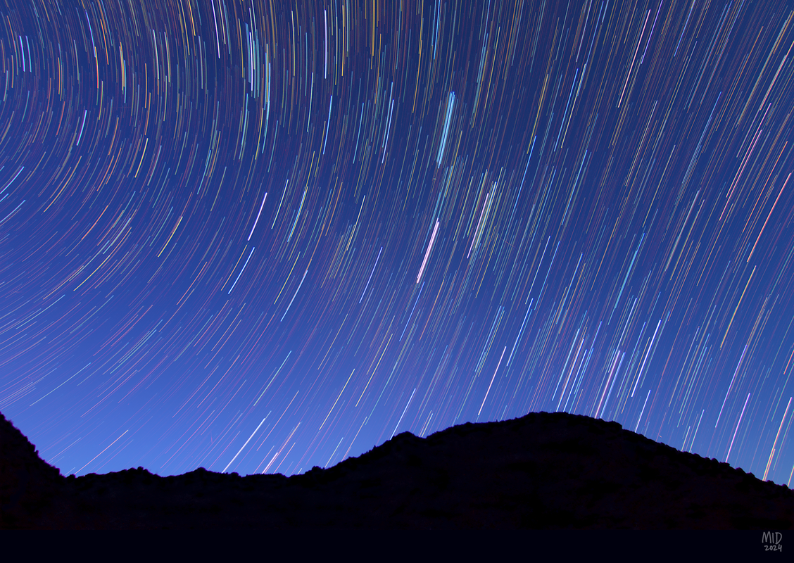 Photograph of many multicolored star trails over the silhouette of a rocky ridge. The sky is pre-dawn blue.