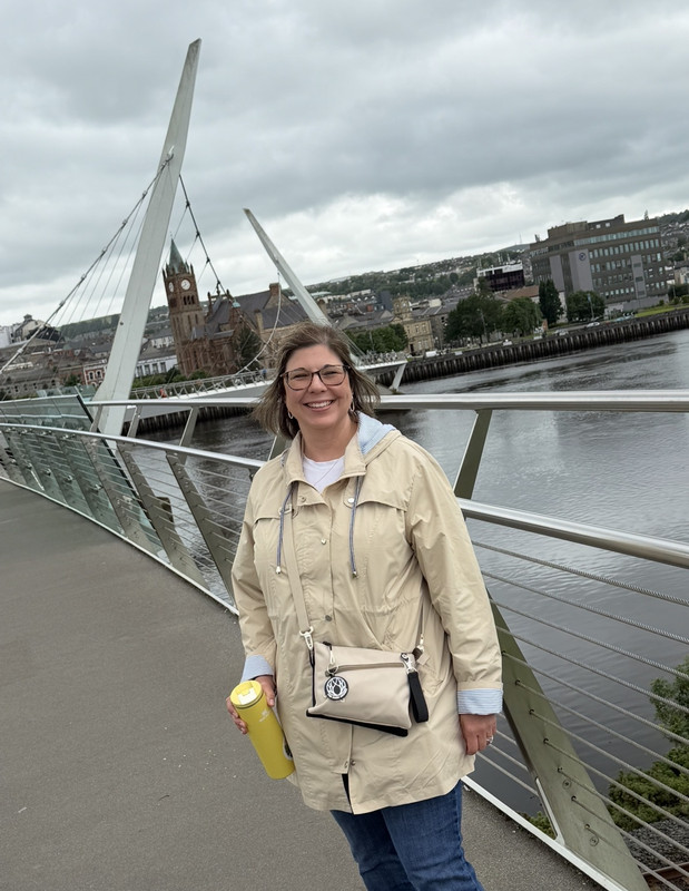 Dr. Stefanie Sorbet, a smiling woman with glasses and shoulder-length brown hair, standing outdoors on a pedestrian suspension bridge. She is wearing a beige jacket over jeans, holding a yellow travel tumbler, and wearing a light-colored crossbody bag, with a river, city architecture, and an overcast sky in the background.