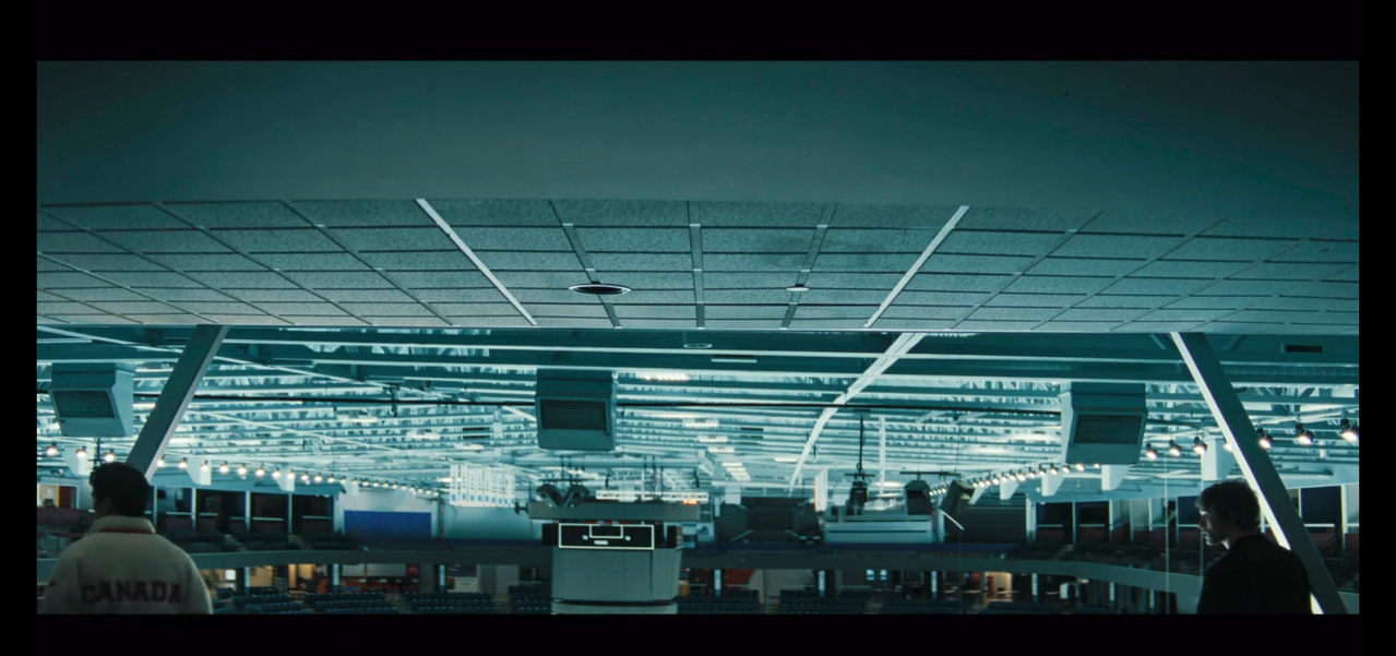 a wide shot showing shane and ilya on extreme ends of the image, shane on the left and ilya on the right. they are small in the composition with the rafters of a skating rink filling the rest of the screen. shane is walking offscreen and ilya is watching him go. the word canada is clearly visible on the back of shane's fluffy white jacket. ilya's in a black coat.