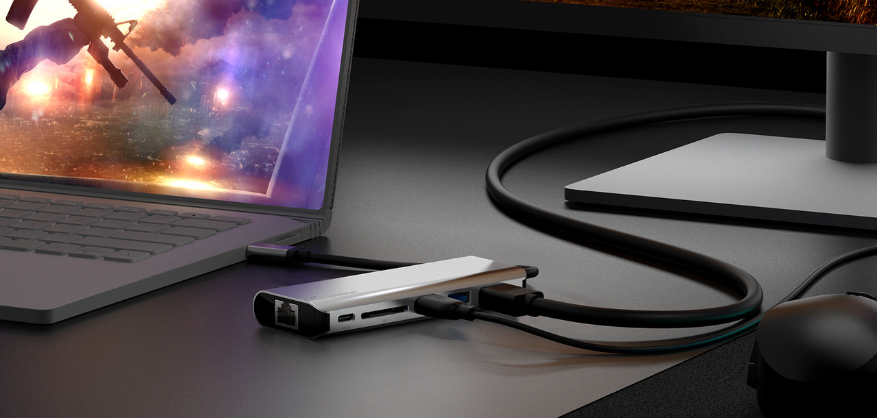 Adaptador multipuerto Belkin conectado a una laptop junto a un monitor y otros periféricos.