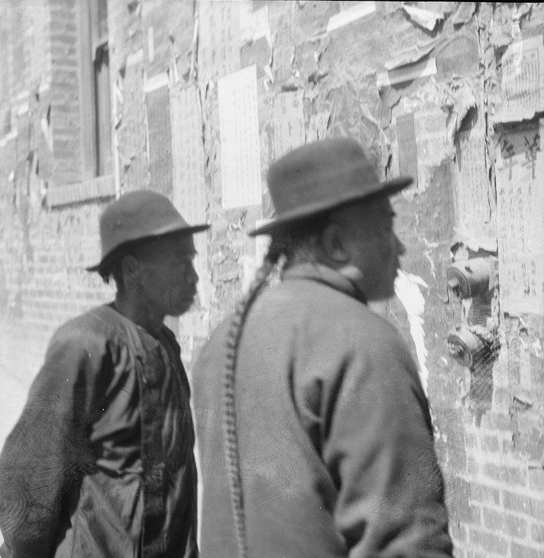 Photograph showing a pair of men in San Francisco's Chinatown reading (in Mandarin) some notices pinned to the wall, circa 1890-1910.