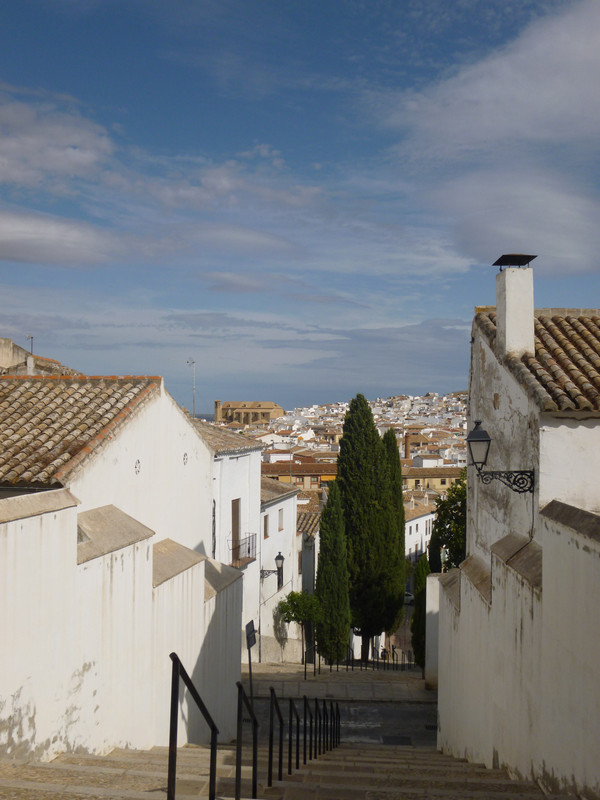 A view down some steps. 
 On the two sides are white painted houses with orange tiled roofs.  In the centre of the stairs is a fir tree.  In the distance is an orange stone church.