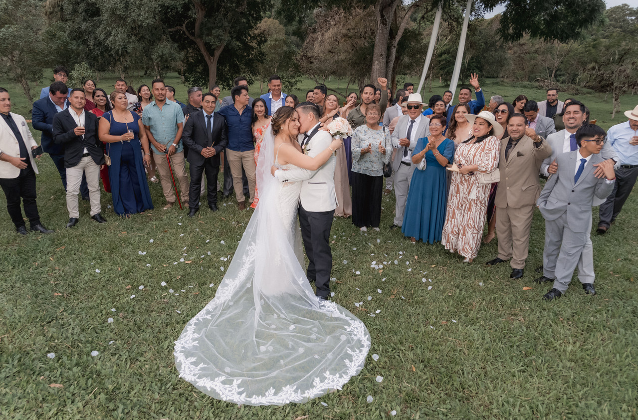 Esposos abrazados con invitados de fondo, vestido al viento