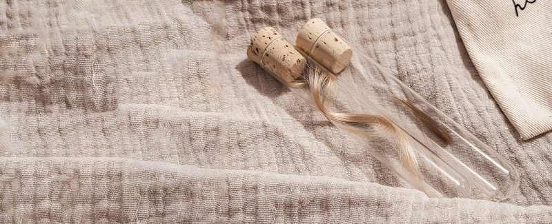 Twee glazen bewaarbuisjes met haarlokjes van een kindje, naast een katoenen bewaarzakje om de eerste haarlokjes als dierbare herinnering te bewaren.