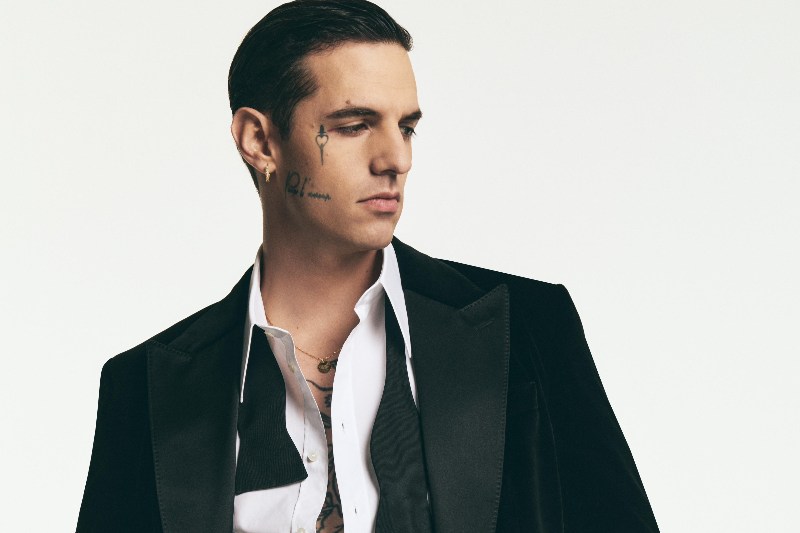 Achille Lauro: Il Compleanno dell'Artista Poliedrico l'11 Luglio Primo piano di Achille Lauro, nato l'11 luglio 1990, con taglio di capelli all'indietro, tatuaggi sul viso e collo, che indossa una camicia bianca sbottonata e una giacca scura, guardando di lato su sfondo bianco.