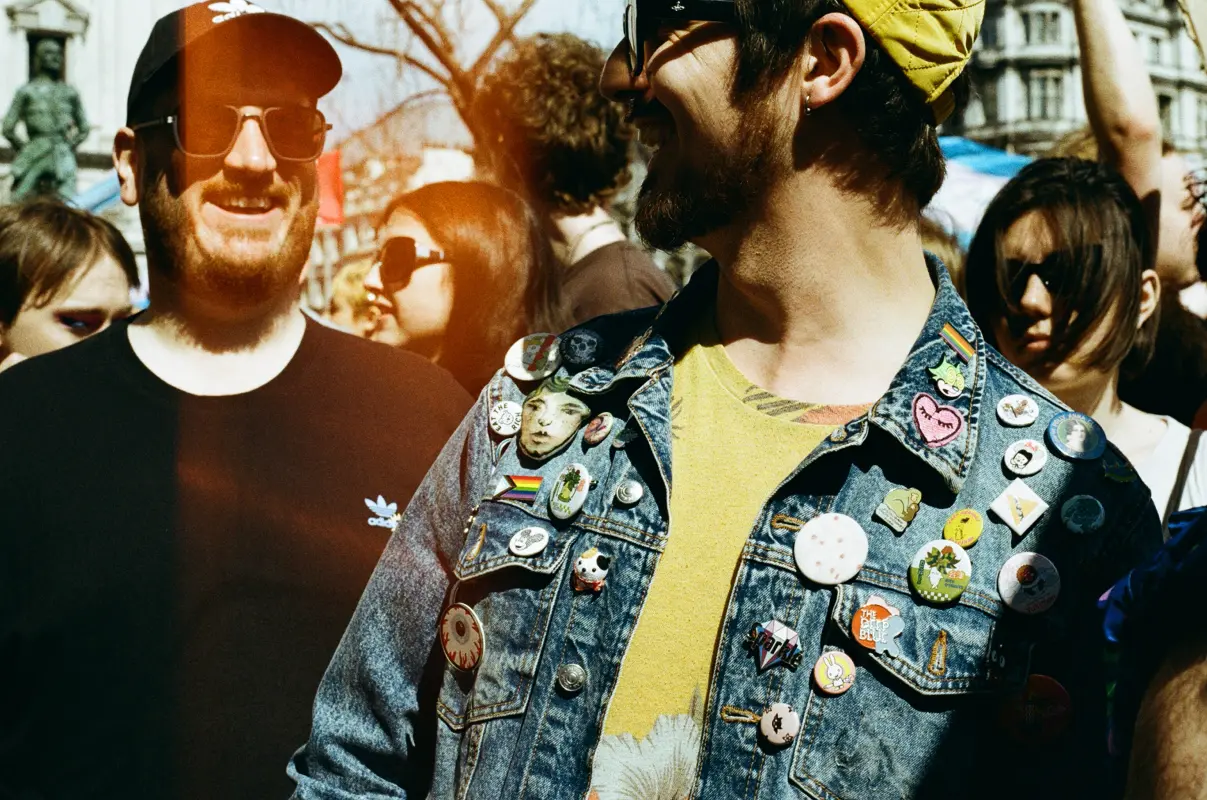two people in sunglasses looking at each other laughing, the one in the front is wearing a jean jacket full of cute pins and badges.
