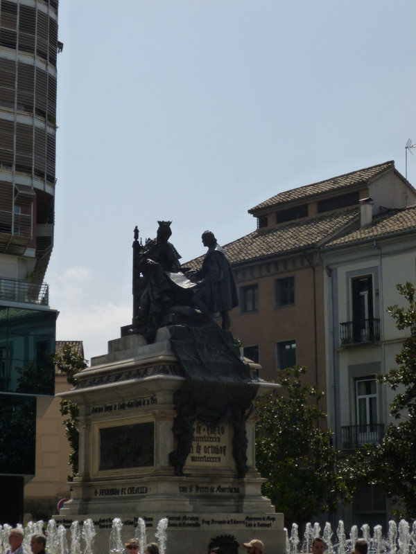 Statue of Christopher Colombus kneeling in front of Isabella the Catholic.  The plinth is an off white stone, the figures are in bronze.  Isabella dress cascades down the front of the plinth in ripples.