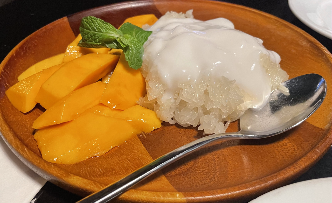 a photo of a wooden plate of mango sticky rice with a spoon laying across it and mint on top.