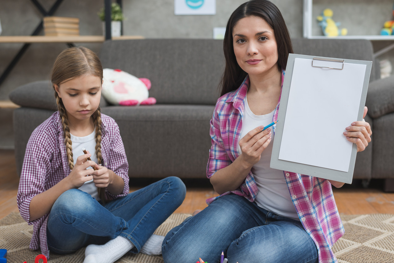 professional-female-psychologist-sitting-with-girl-carpet-showing-white-paper-clipboard.jpg