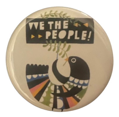 a white pin with a colorful & stylized bird on it that's holding olive branches and a protest sign that says 'WE THE PEOPLE!'