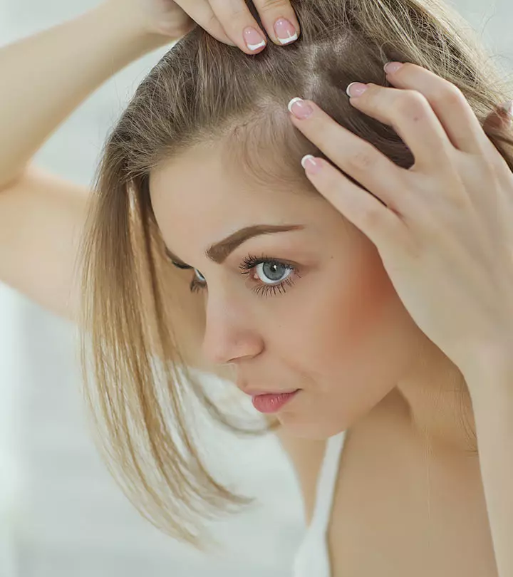 A blonde woman looks at the roots of her hair, which has started to fall out, in the mirror