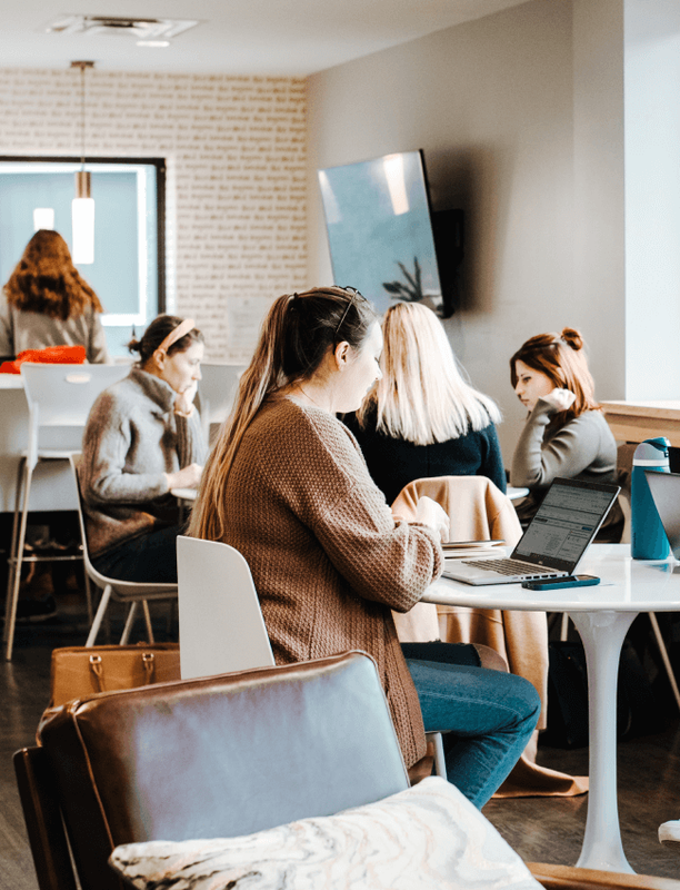 Multiple people working individually on laptops in a bustling coworking common area.