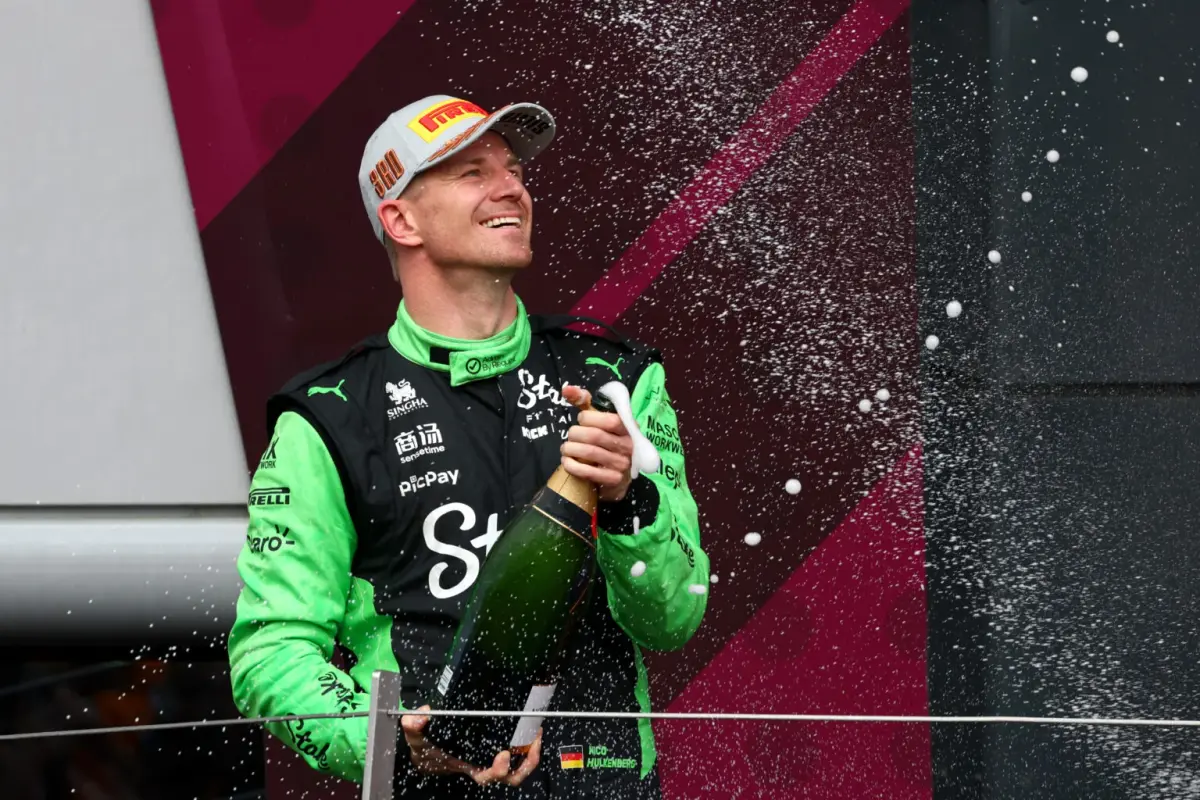 Picture of Nico Hulkenberg (tall, skinny, white, blond) on the podium at the British Grand Prix.  He is wearing a white baseball cap and his black and green Kick Sauber racing overalls.  He is holding a bottle of champagne which is spraying foam.  He has a very relieved smile.