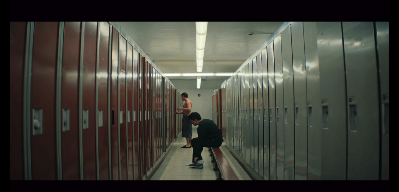 shane and ilya in the locker room. shane sits on a bench in the middle of the frame, dressed in all black except for his socks. he's in a long-sleeved black shirt and pants. ilya stands at his locker in the background, naked except for the gray-green towel around his waist and his black slides.