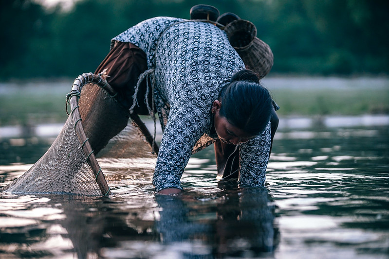Een Nepalese vrouw staat in een rivier in Nepal. Ze buigt voorover in het water en vangt vis met haar handen en een net.