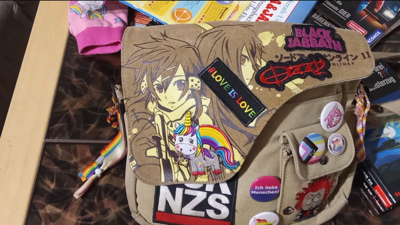 A brown Sword Art Online messenger bag with multiple pins and patches visible on the front flap of the bag which is designed to have a lopsided shape to expose a side pocket even when closed 4 patches are visible black sabbath ozzy osbourne #love is love and a unicorn with a rainbow. above the side pocket is a pin with a pink haired anime girl's face with the phrase Map Rights. On the side pocket top flap are two pins one of them being an edited version of the progress flag to have the pedophile flag instead of the rainbow the other having a rainbow silhouette of what seems to be a mouse? It has text but it's too blurry to read in this photo. On the pocket itself is a patch of some weird vampire looking south park character. next to the side patch are two more pins, one with the bisexual flag and the text Ich liebe Menschen! meaning I love men! the other pin appearing to be a rainbow silhouette of germany  and a FCK NZS patch. The bag also has two fabric hairties attached on the left side as accessories one being a rainbow the other being the pedophile flag. on the right is a hand woven lesbian pride bracelet attached in a similar fashion.