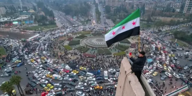 صورة توضيحية لمشهد احتفالي في ساحة الأمويين بدمشق، حيث يرفع أحد الأشخاص العلم السوري الجديد من أعلى نقطة في الساحة المزدحمة بالسيارات والمحتشدين، في مشهد يعكس التحولات السياسية الكبرى في سوريا وعودة الأمل بمسار سياسي جديد.