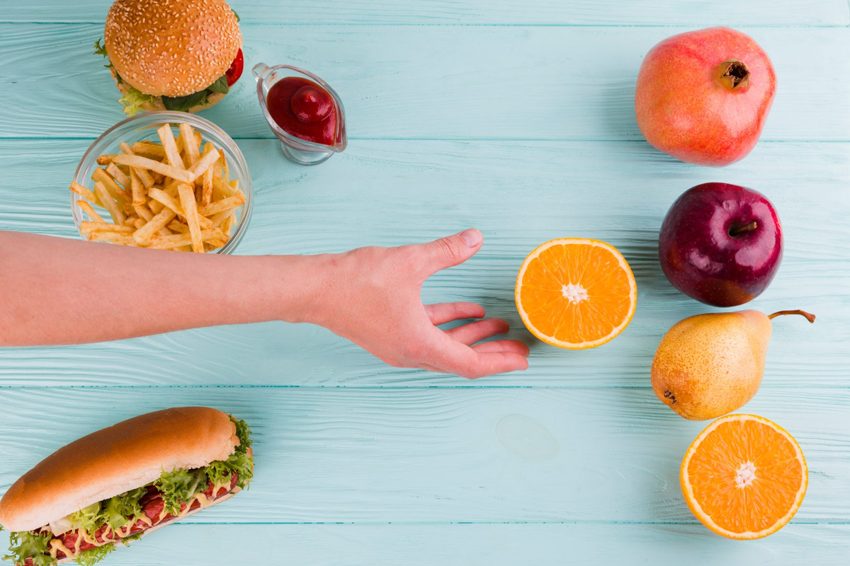 Mano che sceglie frutta fresca invece di hamburger e patatine fritte su sfondo azzurro.